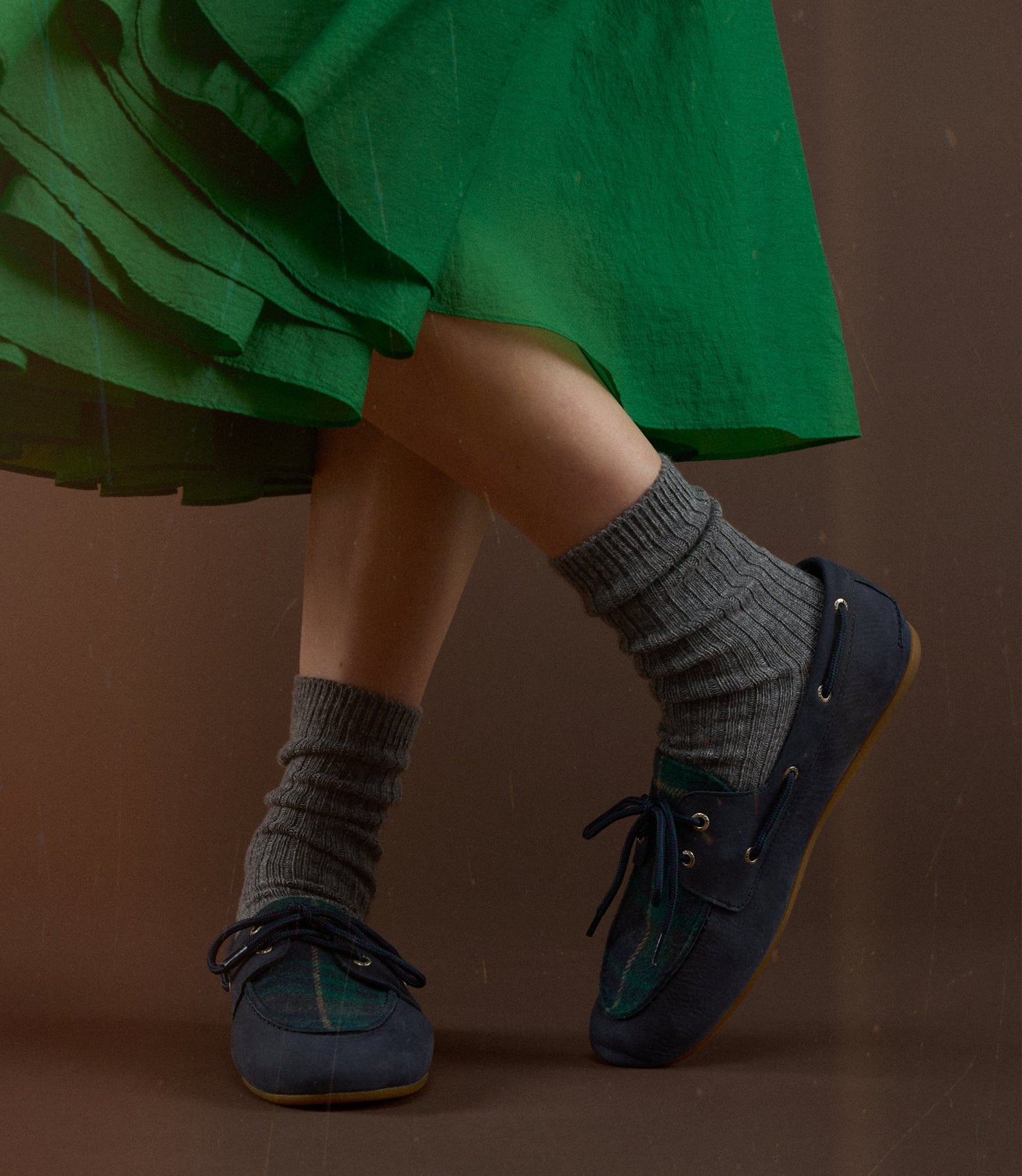 Close-up of a person in a green pleated skirt and grey wool socks wearing navy Sperry boat shoes with green accent lacing.