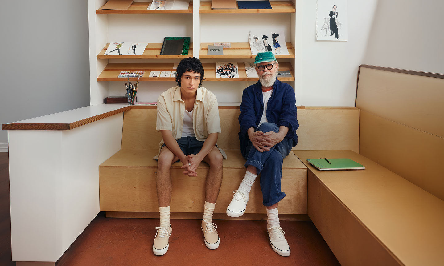 Two men of different generations seated on a wooden bench in a studio setting, both wearing white Sperry CVO canvas sneakers with crew socks, surrounded by sketchbooks and art supplies on shelves behind them.