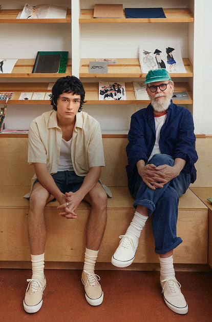 Two men of different generations seated on a wooden bench in a studio setting, both wearing white Sperry CVO canvas sneakers with crew socks, surrounded by sketchbooks and art supplies on shelves behind them.