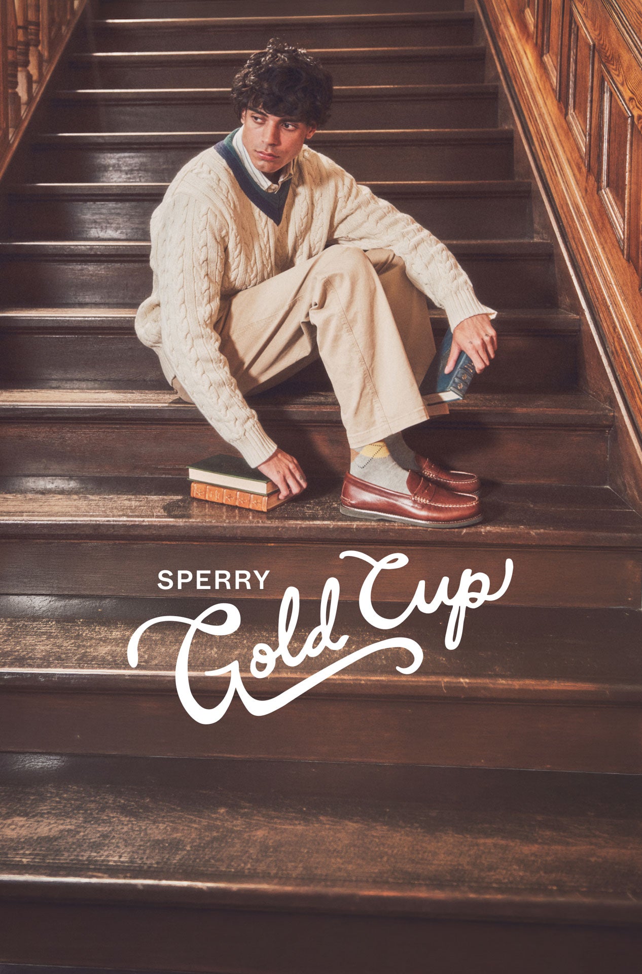 A man sitting on a brown staircase wearing brown loafers and holding books