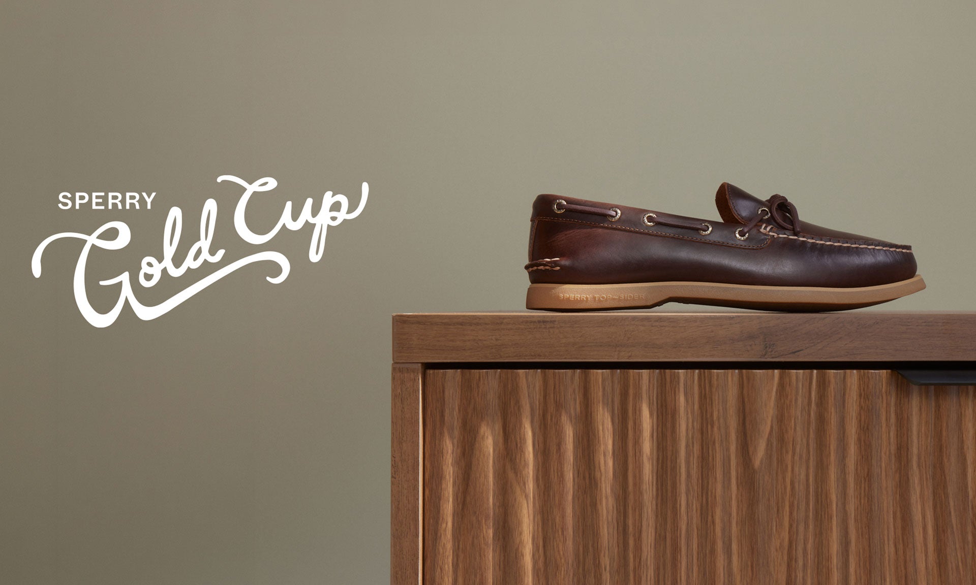 Dark brown Sperry Gold Cup leather boat shoe with a gum sole displayed on top of a walnut wood cabinet against a warm grey background.