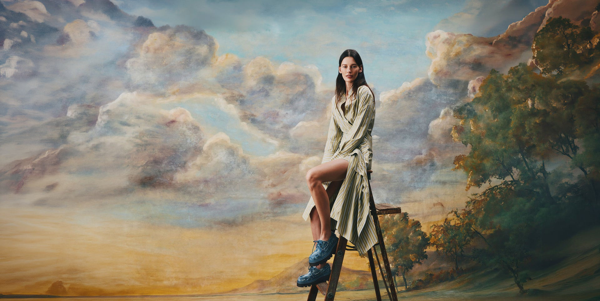 Woman in a flowing striped dress seated on a wooden ladder wearing blue leather Sperry x MONSE lug boat shoes, set against a classical painted landscape backdrop.