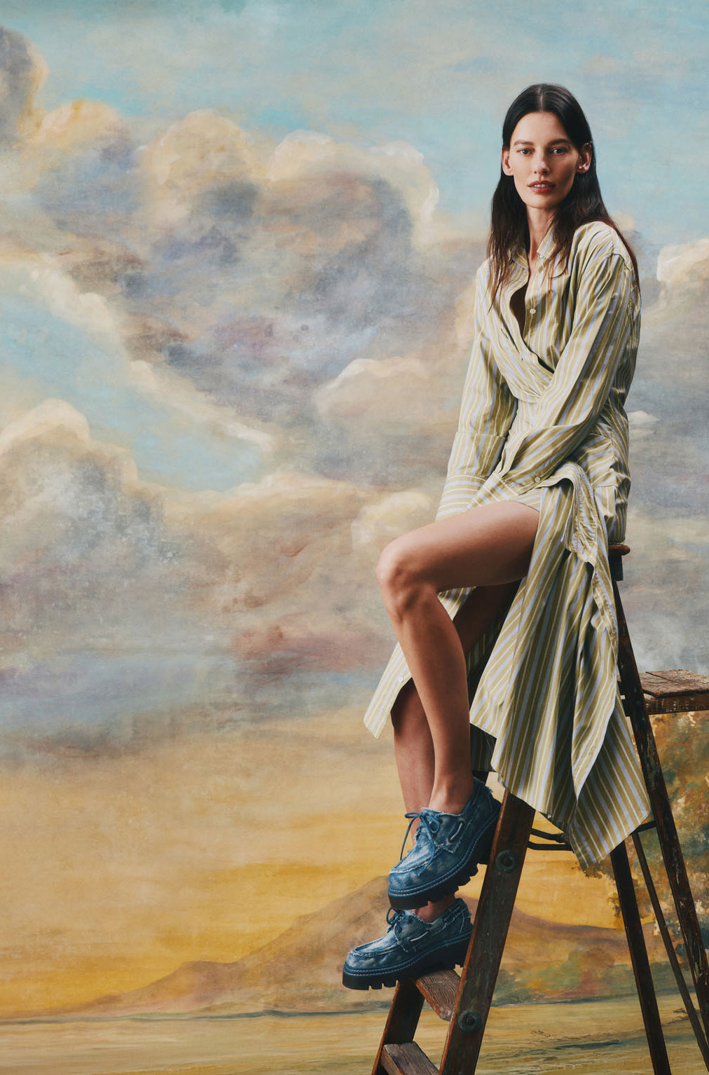 Woman in a flowing striped dress seated on a wooden ladder wearing blue leather Sperry x MONSE lug boat shoes, set against a classical painted landscape backdrop.