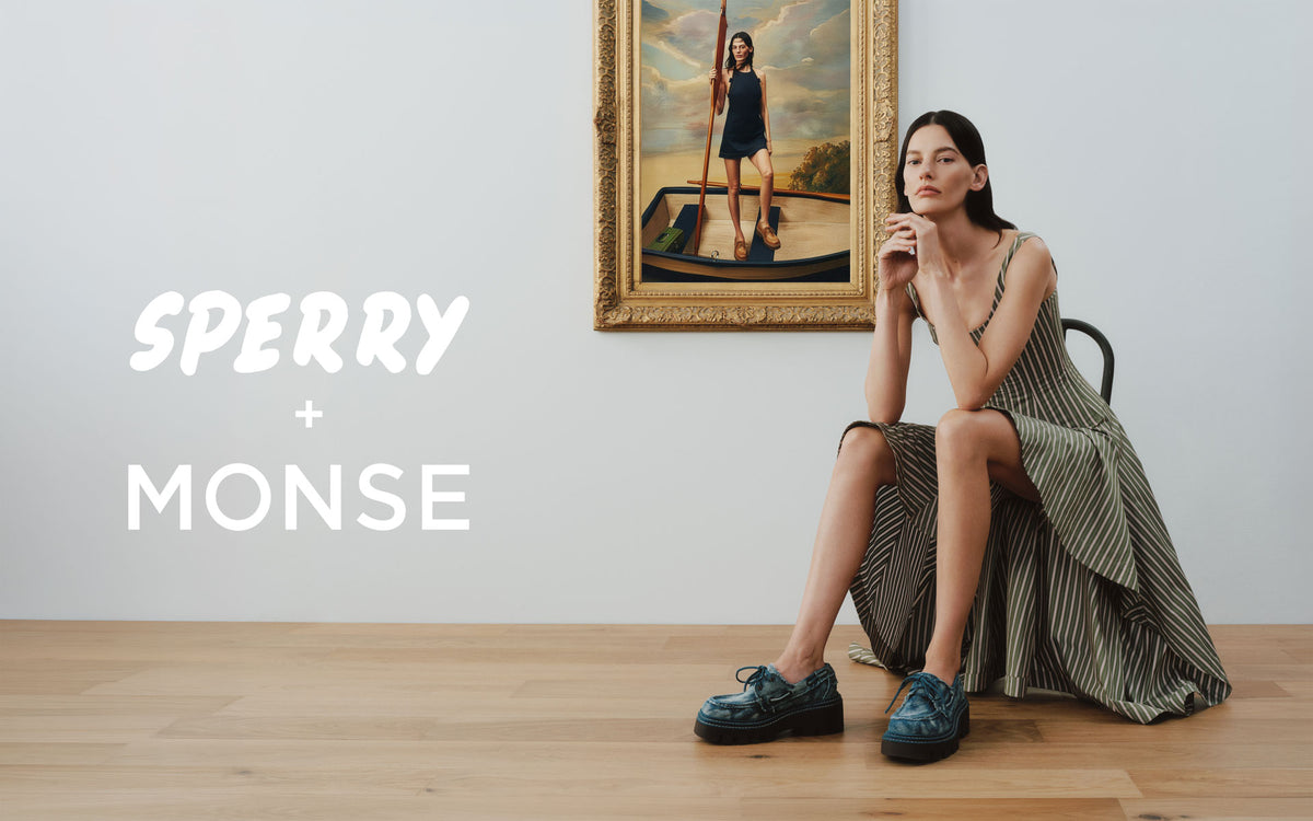 Woman in a flowing striped dress seated in a gallery space wearing blue denim Sperry x MONSE lug boat shoes, with a classical painting of a figure in a boat hanging on the wall behind her.
