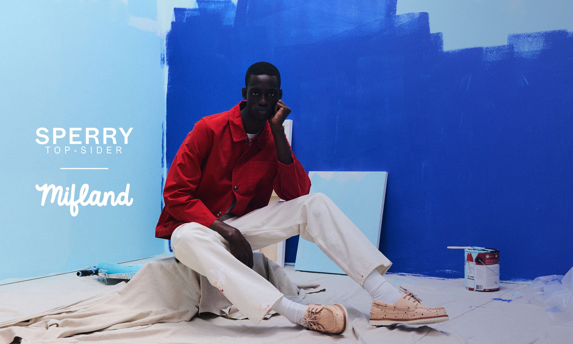 Homme assis sur un plancher d'atelier éclaboussé de peinture, portant une veste rouge, un pantalon crème et des chaussures bateau Sperry x Mifland beige, entouré de rouleaux et de pots de peinture devant un mur peint en bleu vif