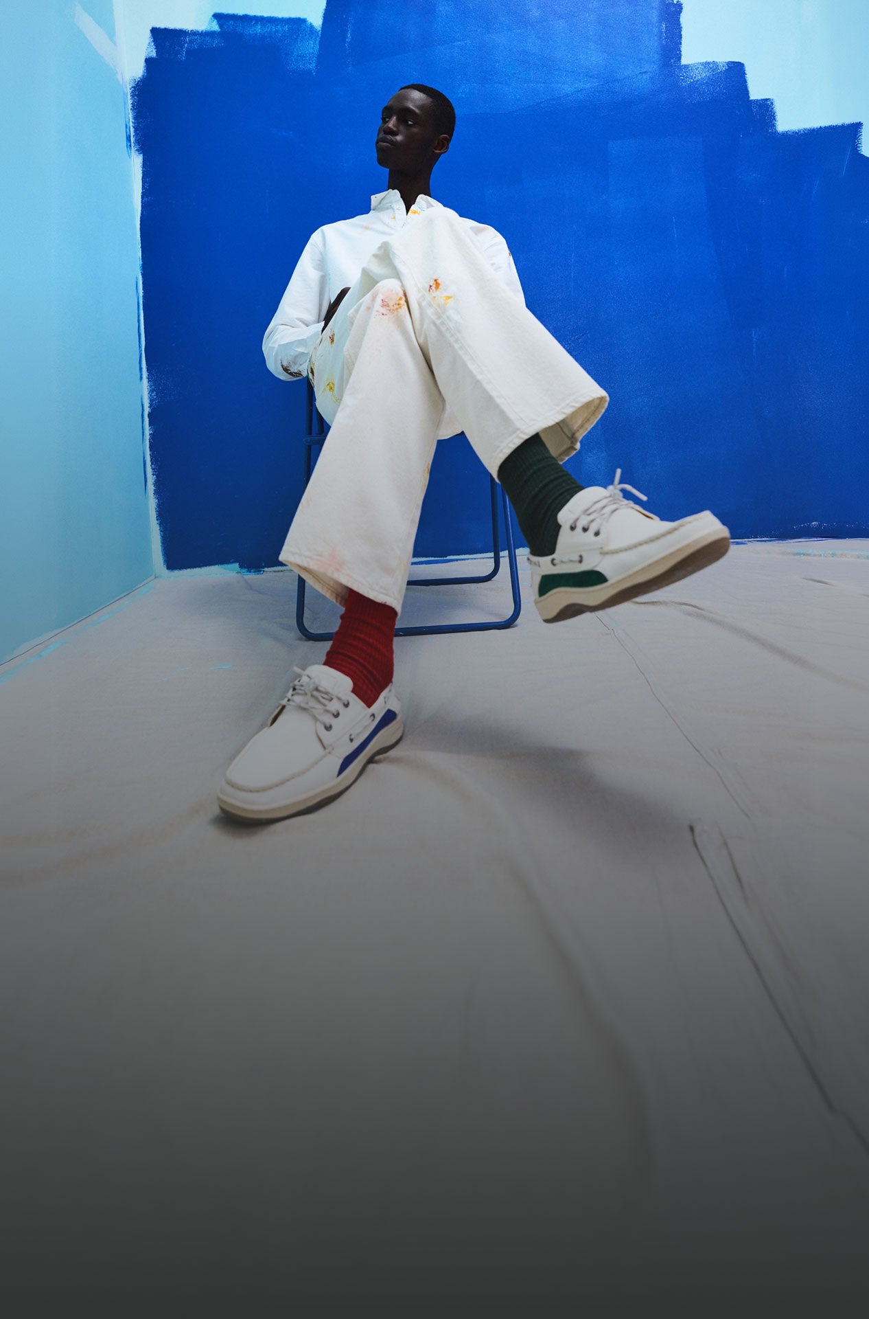 Homme assis sur un plancher d'atelier éclaboussé de peinture, portant une veste rouge, un pantalon crème et des chaussures bateau Sperry x Mifland beige, entouré de rouleaux et de pots de peinture devant un mur peint en bleu vif
