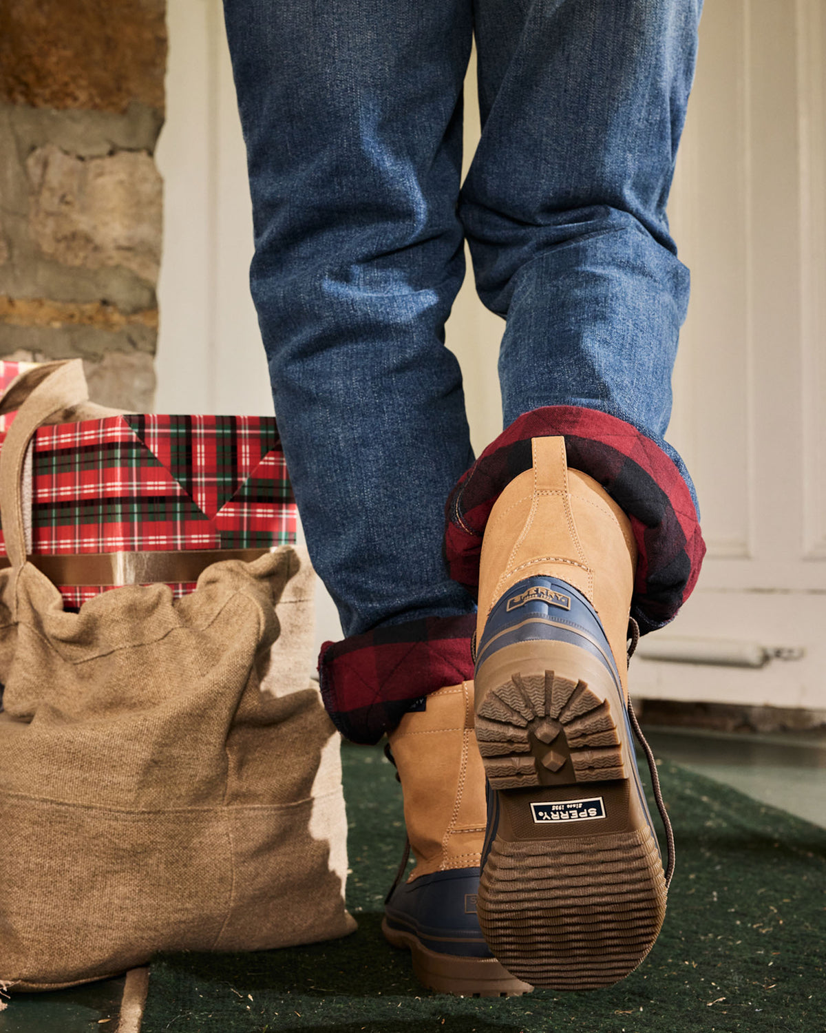 Homme en jean qui avance, portant des bottes canard brun clair et bleu marine, avec un sac de jute et une boîte-cadeau à carreaux à côté de lui.