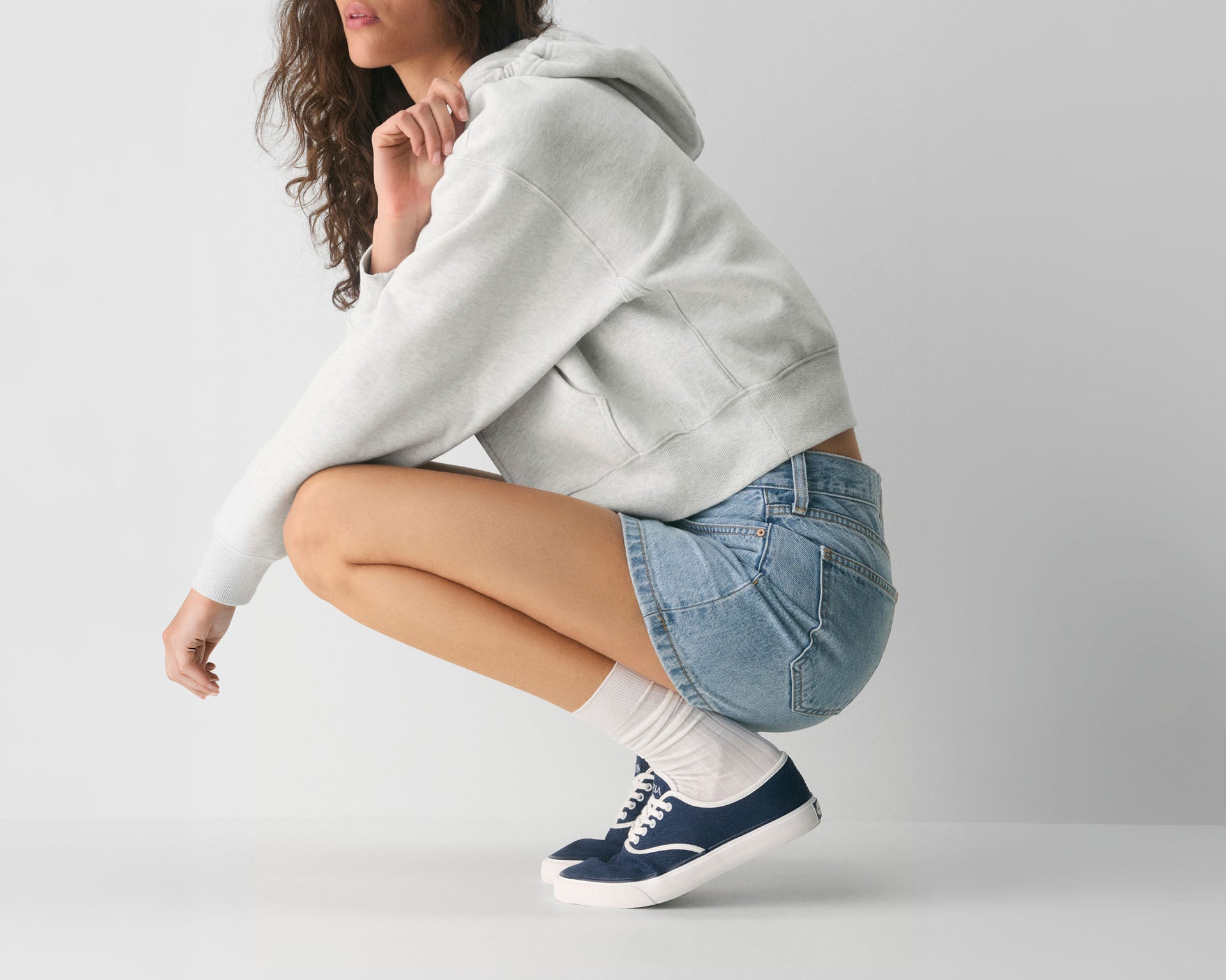 Woman in a grey hoodie and denim shorts crouching, wearing navy Sperry canvas sneakers with white crew socks against a light grey background.