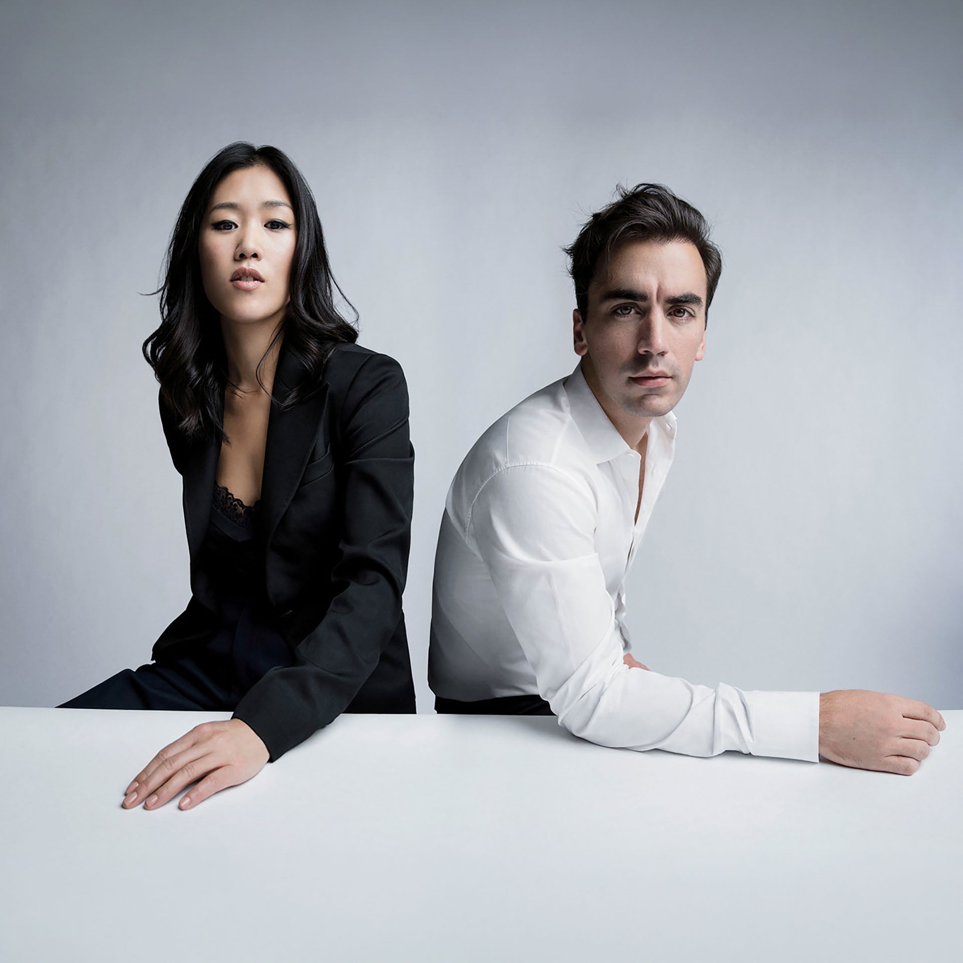 Two fashion designers posed at a white table against a grey background — a woman in a black blazer and a man in a white dress shirt — likely the creative directors behind the Sperry x MONSE collaboration.