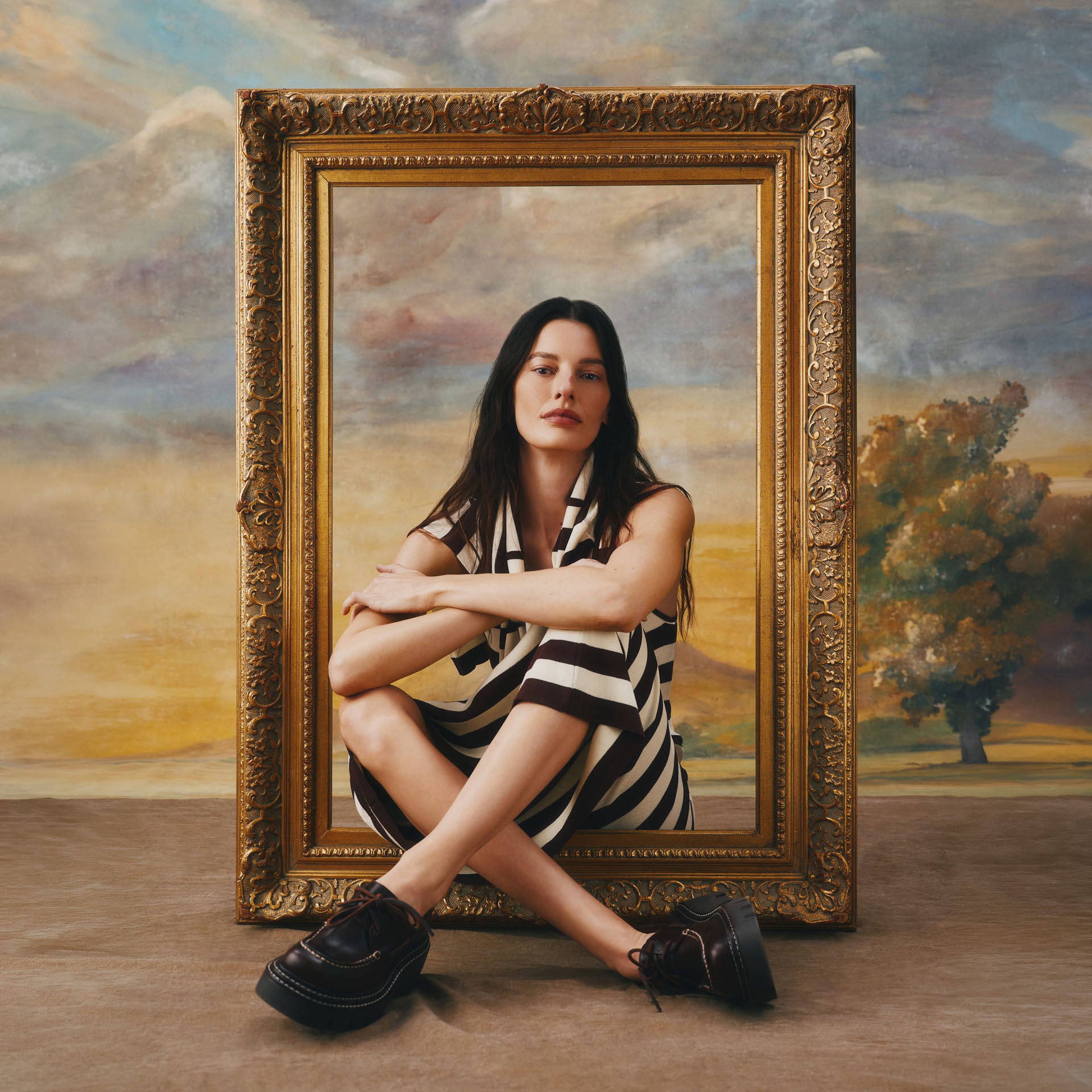 Woman in a black and white striped dress seated cross-legged in front of an ornate gold picture frame, wearing black platform Sperry x MONSE lug boat shoes, against a classical painted landscape backdrop.