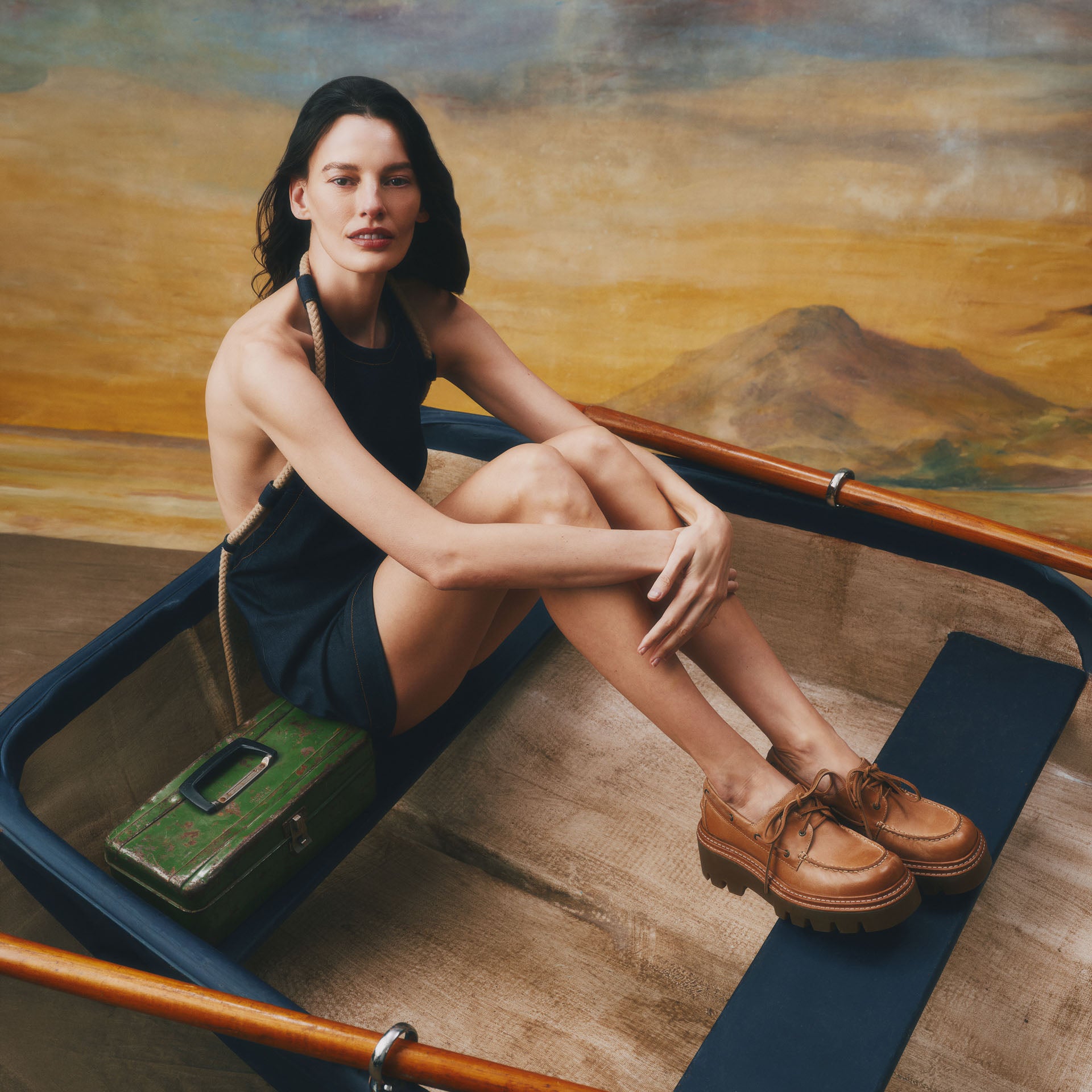 Woman in a black halter dress seated inside a small wooden boat wearing tan leather Sperry x MONSE lug boat shoes, with a vintage green toolbox beside her and a classical painted landscape in the background.