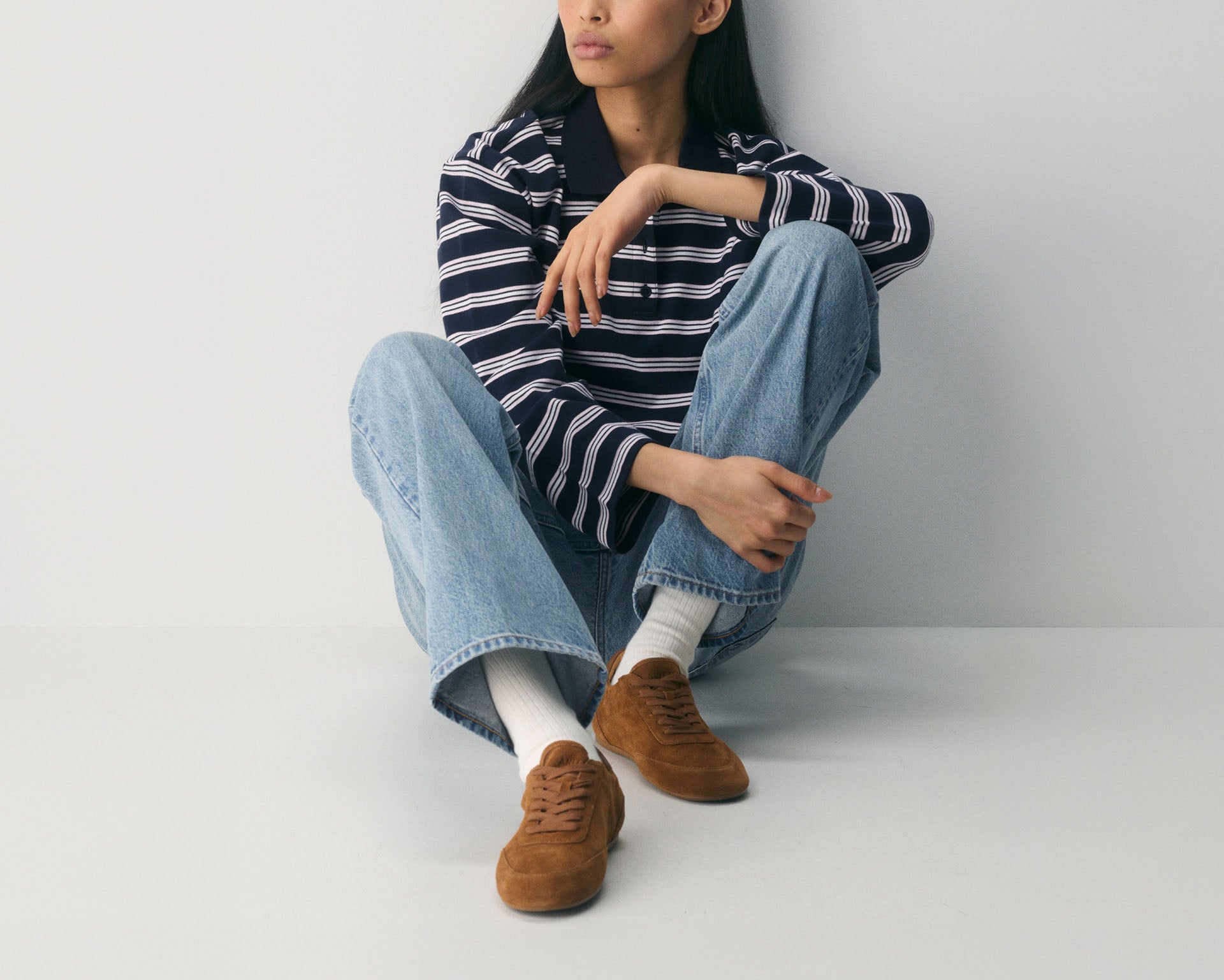 Woman in a navy striped long-sleeve shirt and wide-leg jeans seated on the floor, wearing brown suede Sperry sneakers with a gum sole and white crew socks.