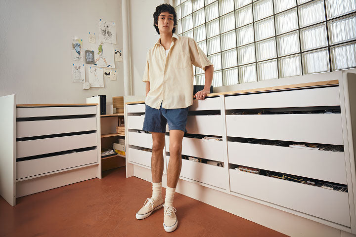 Young man in a cream short-sleeve shirt and blue shorts standing in a bright studio space, wearing white Sperry CVO canvas sneakers with crew socks.