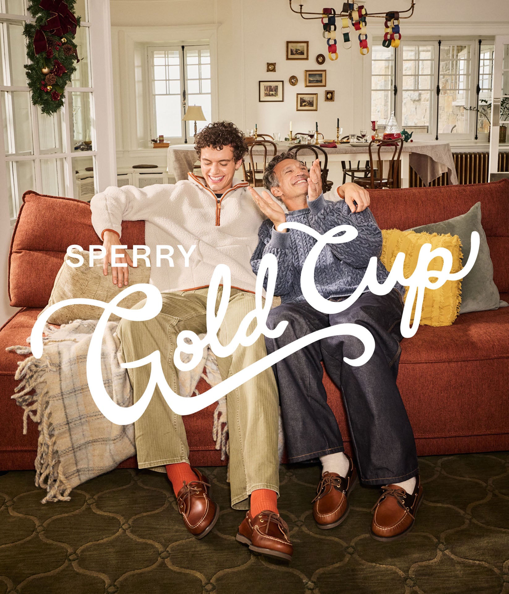 Dad and son laughing together on an orange sofa in a holiday-decorated living room, wearing brown leather Sperry Gold Cup boat shoes.