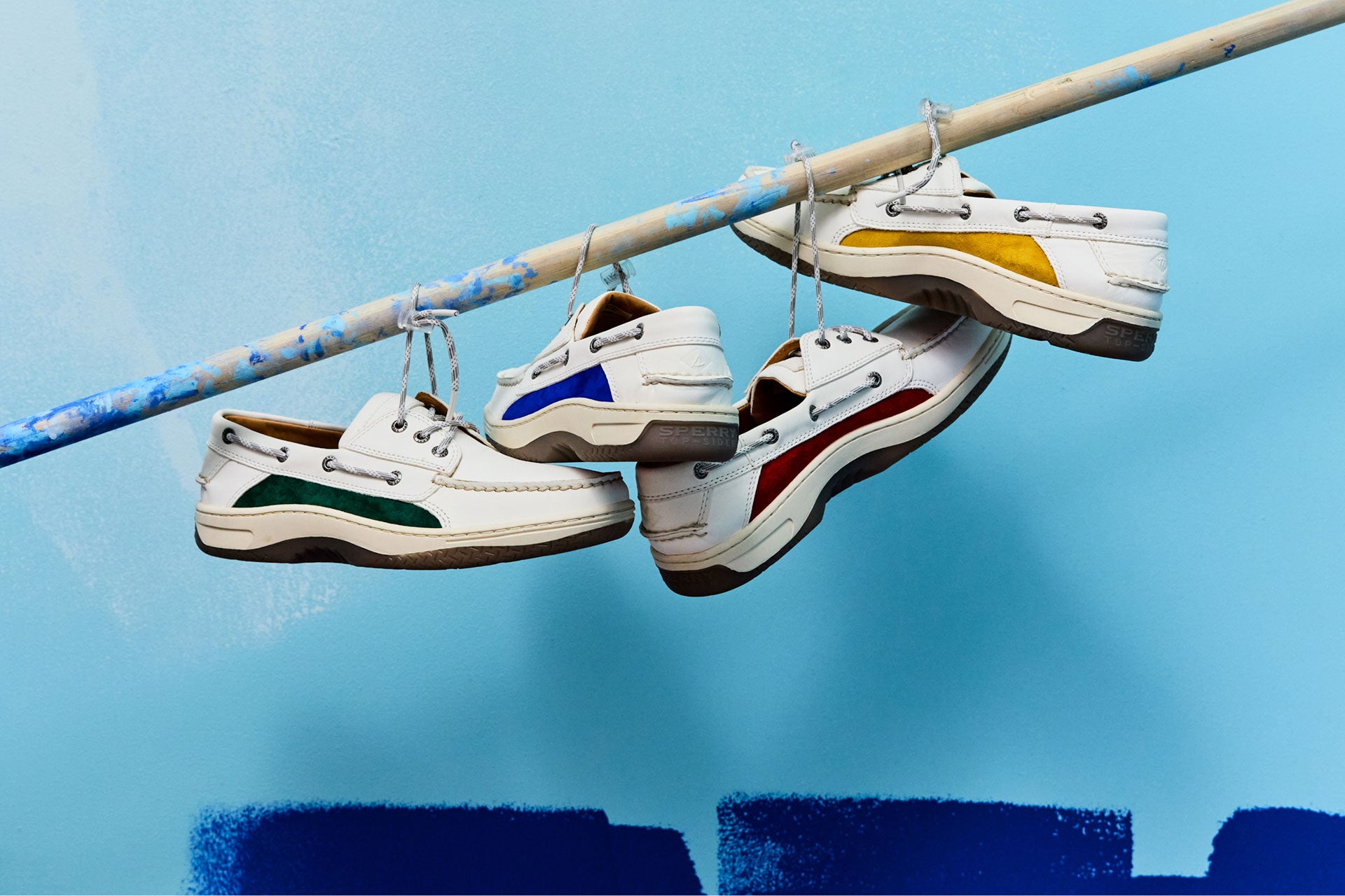 Trois paires de chaussures bateau Sperry x Mifland Billfish en cuir blanc avec des panneaux color-block aux couleurs vives — jaune, bleu, vert et rouge — suspendues par leurs lacets à une tige éclaboussée de peinture sur fond bleu clair.