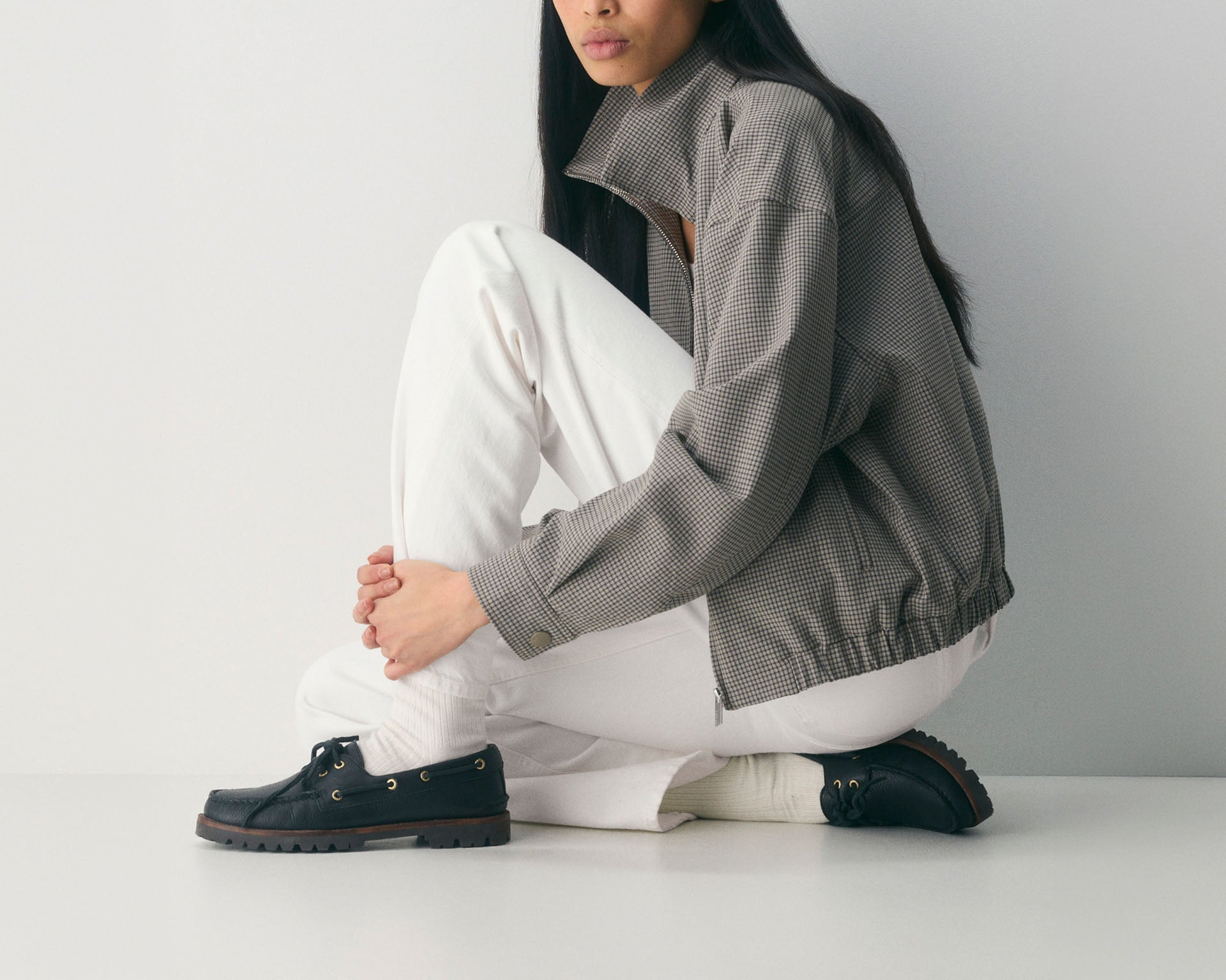 Woman in a grey houndstooth jacket and white jeans seated on the floor, wearing dark navy leather Sperry boat shoes with a chunky lug sole.