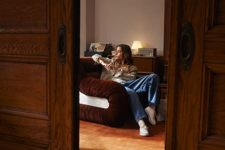 Woman in wide-leg jeans and a metallic top relaxing on a deep red velvet sofa in a warm vintage room, wearing white Sperry platform sneakers, with vinyl records and a retro lamp in the background.