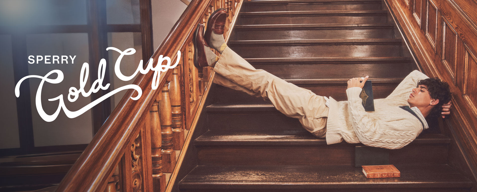 A man lying down on a brown staircase reading a book and wearing brown loafers