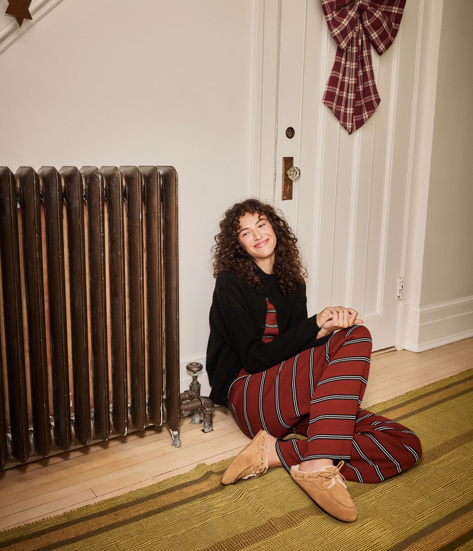 Woman with curly hair sitting on the floor by a radiator in striped red pyjama pants and a black sweater, wearing tan suede slippers.
