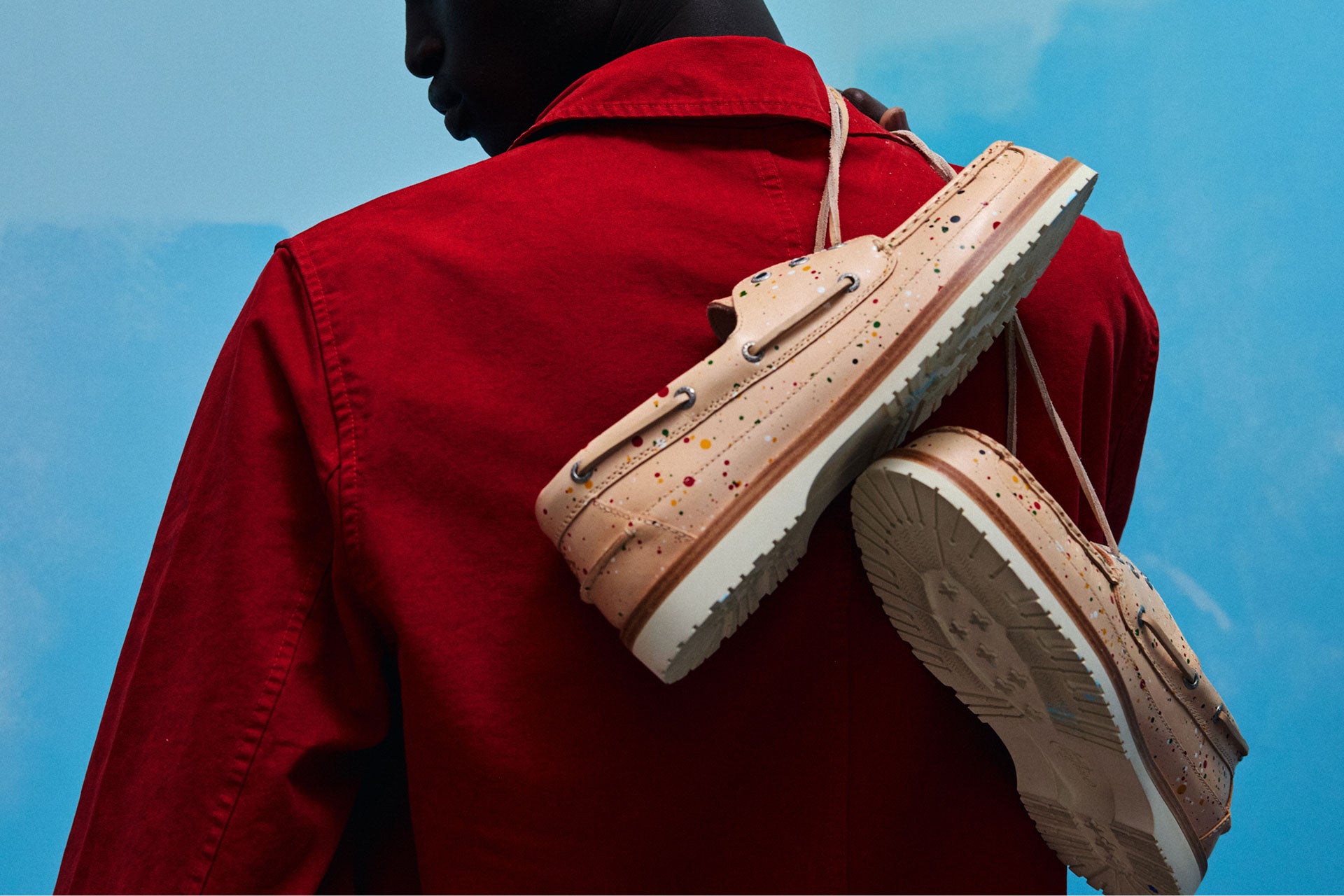 Gros plan d'une personne en veste rouge tenant la chaussure bateau Sperry x Mifland 3-Eye Mini Lug par-dessus son épaule, montrant la semelle en cuir beige avec des détails d'éclaboussures de peinture colorées.