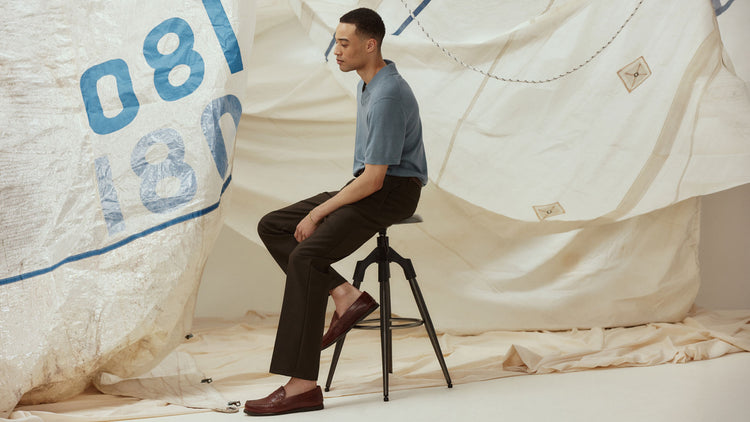 Man in a tropical print shirt and dark trousers seated on a green velvet chair wearing cognac leather penny loafers, alongside a close-up of the shoe with grey dress socks.