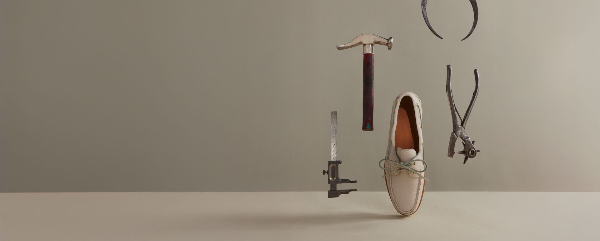 White Sperry Gold Cup leather boat shoe suspended in mid-air surrounded by floating cobbler tools — a hammer, pliers, caliper, and lasting pincers — against a grey background, highlighting the handcrafted construction process.