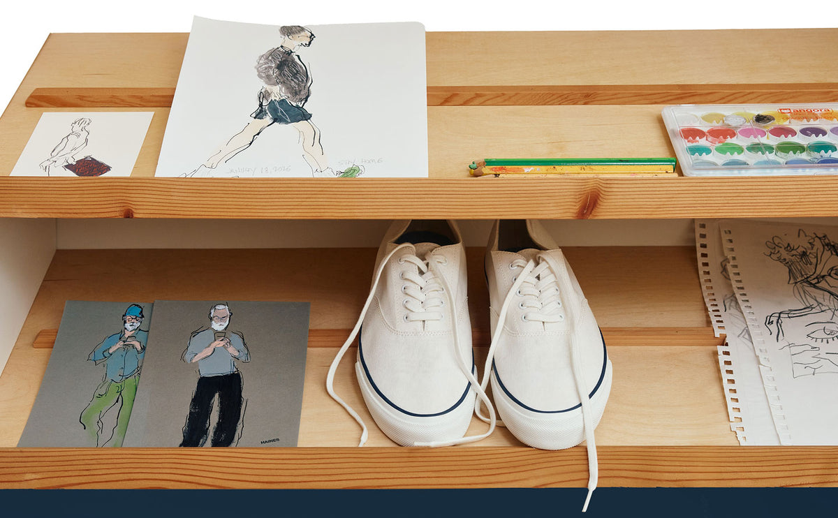 Pair of white Sperry CVO canvas sneakers with navy accent stitching resting on a wooden shelf alongside fashion illustrations by Richard Haines, watercolor paints, pencils, and sketch notebooks.