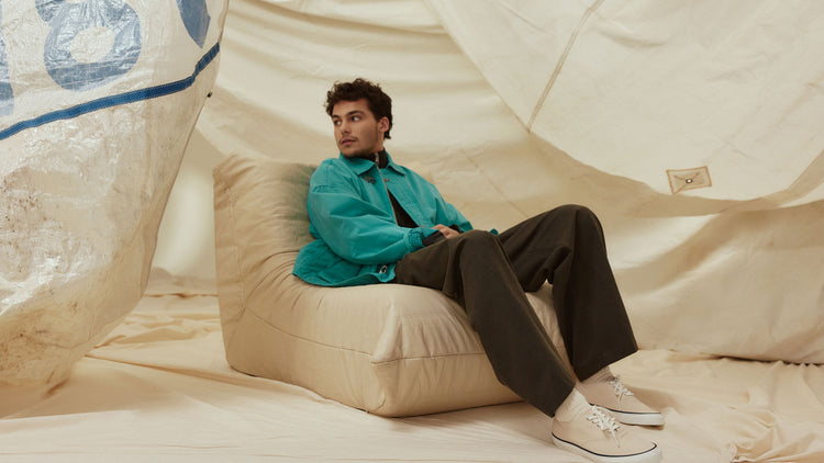 Man in a black varsity jacket and trousers seated on a chrome chair wearing black Sperry leather sneakers with red socks, alongside a close-up of the shoe.