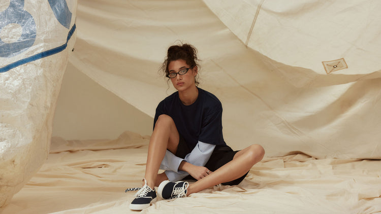 Woman in a pink mini skirt seated on a green velvet chair wearing white Sperry high-top sneakers with floral embroidery and ruffle laces, alongside a close-up of the shoe detail.