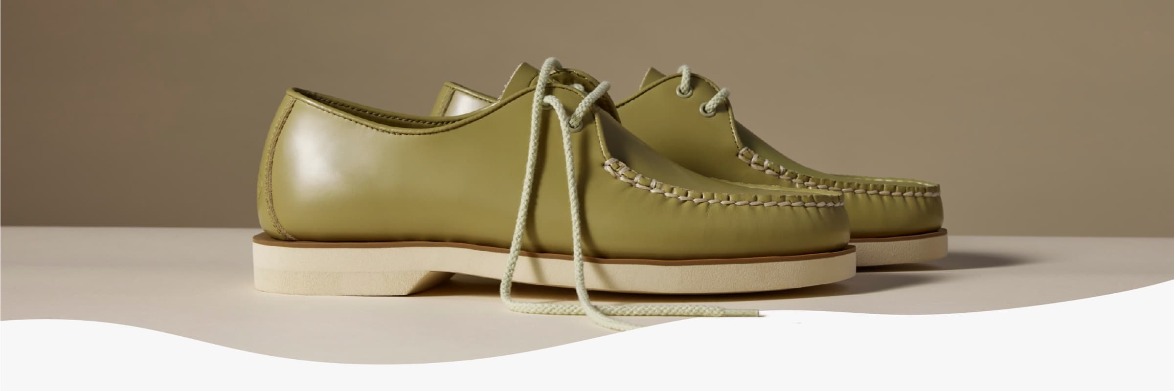 Pair of sage green leather moccasin-style shoes with rope lacing, hand-stitched welt detail, and a cream rubber sole, displayed on a neutral background.