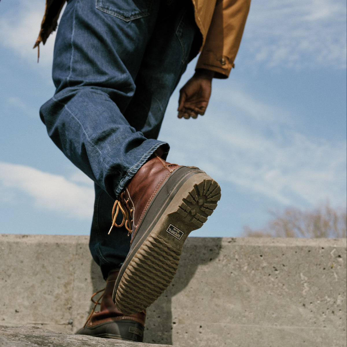 Person in dark jeans stepping up a concrete ledge, showing the ridged olive rubber sole of a brown leather lace-up boot.