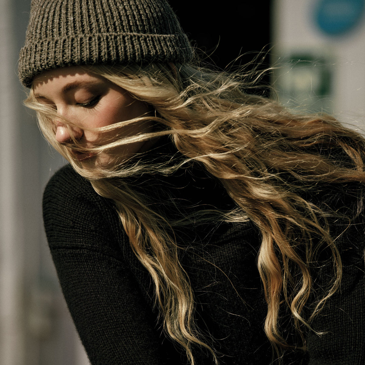 Blonde woman in a dark green knit beanie and black top, eyes closed with her long wavy hair blowing across her face.
