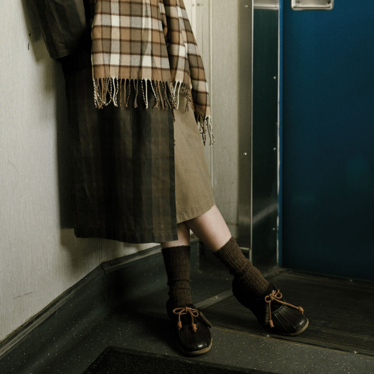 Person in a beige skirt, plaid fringed scarf and brown wool socks, wearing black rubber duck boots with leather laces.
