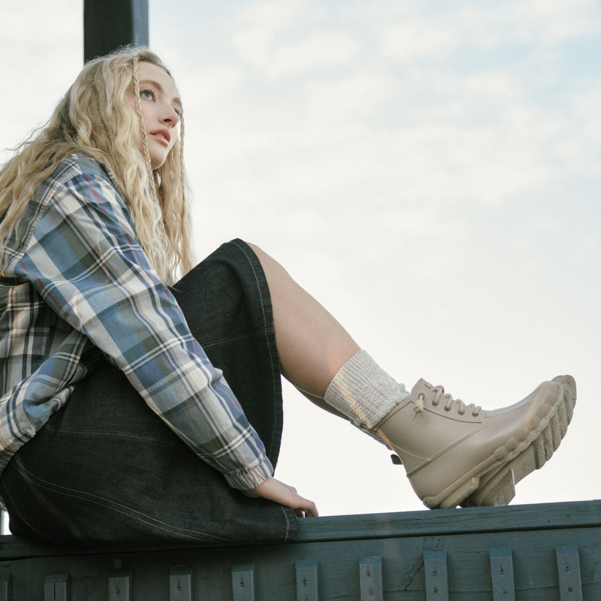 Woman with long blonde hair sitting on a wooden ledge against a pale sky, wearing a plaid shirt, dark skirt and cream lace-up boots.