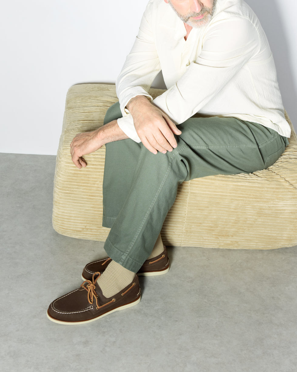 Man seated in olive chinos wearing dark brown Sperry leather boat shoes, alongside a flat-lay of three styles: a dark brown suede boat shoe, a white canvas lace-up sneaker, and a burgundy leather moccasin, presented as Father's Day gift options.