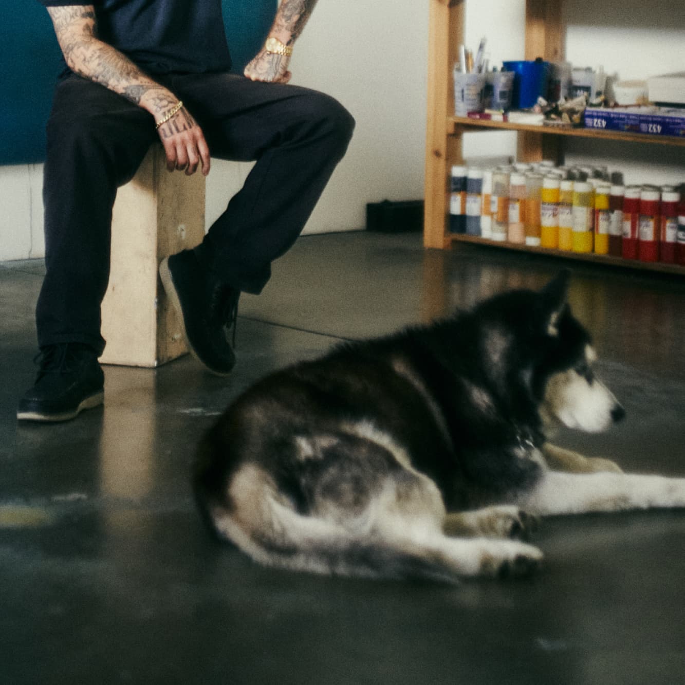 Tattooed man in black outfit seated on a concrete block in an art studio wearing black Sperry sneakers, with a husky dog resting on the floor beside him and paint supplies on shelves in the background.