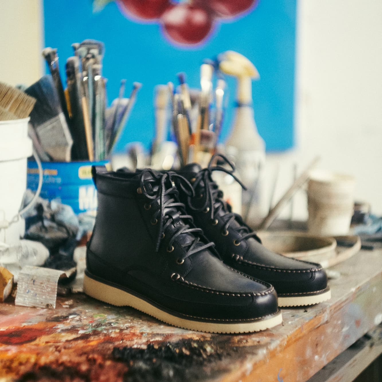 Pair of black leather Sperry moc-toe lace-up boots with a cream sole displayed on a paint-splattered workbench, surrounded by paint brushes and art supplies in an artist's studio.