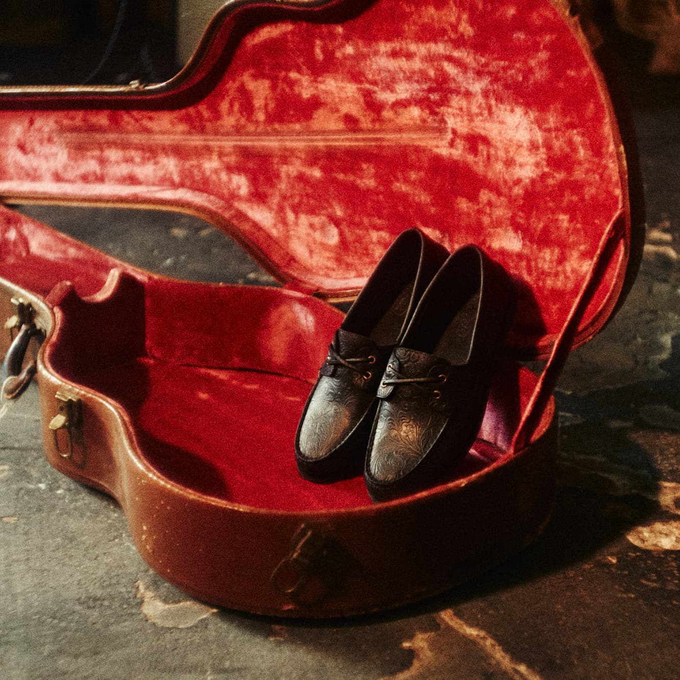 Pair of dark embossed leather Sperry loafers with gold hardware resting inside an open vintage guitar case with red velvet lining.